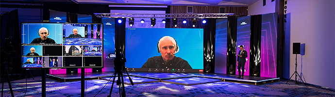 A live event broadcast with a man on a large screen presenting on a topic and several monitors around playing the same broadcast.